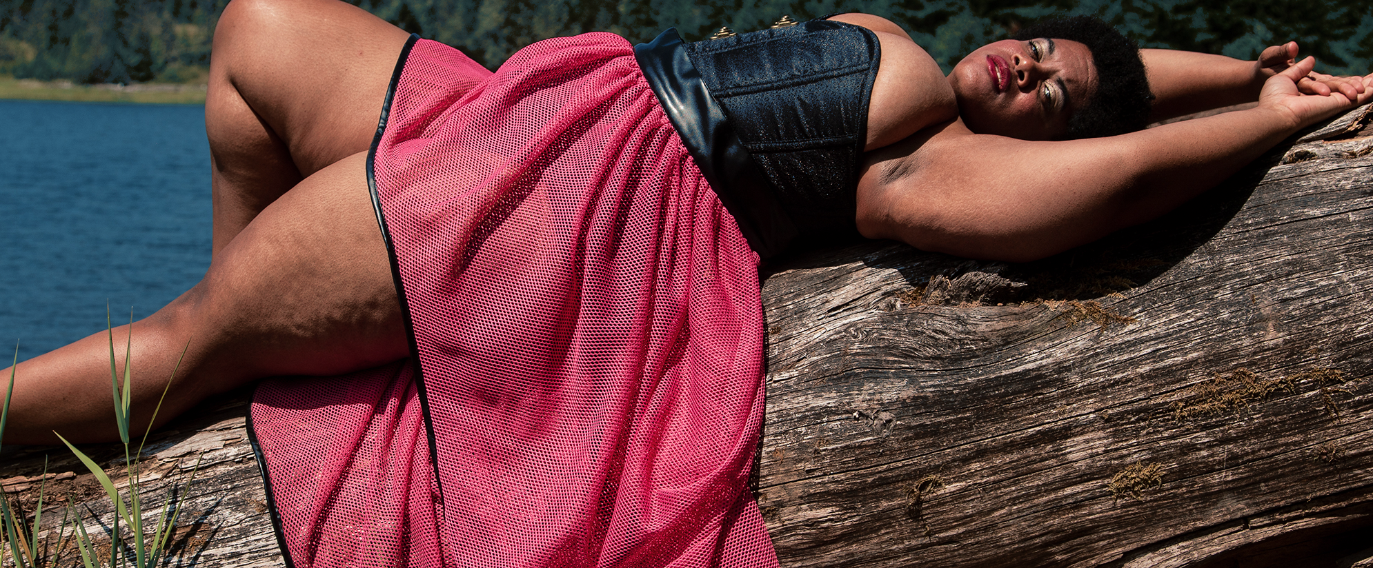 Woman in black corset and pink skirt lying across a tree in front of a lake