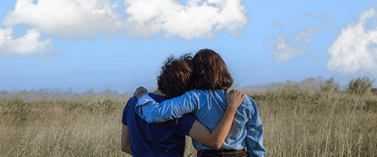 Two people stand with their arms around each other facing away the camera to ward a blue sky with white clouds.